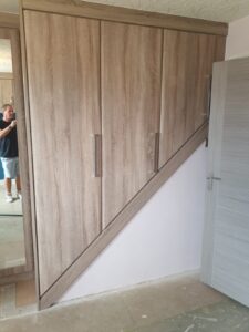 bespoke wardrobes fitted onto a stair bulkhead. The botton of the wardrobes are angled to follow the shape of the bulkhead. The doors are a plain style with a brown-grey oak colour finishe called Sonoma Oak. There are three wardrobes in total.