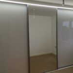 Fitted three door sliding wardrobe with Light Grey coloured glass fitted in the two outer doors and a mirror fitted in the centre door. An LED light strip is fitted above the wardrobe.