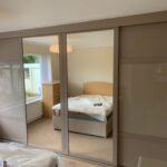 Fitted sliding door wardrobe in the colour stone grey. There are four doors. The two outer doors are fitted with stone grey panels and coloured glass. The two centre doors are fitted with full length mirrors.