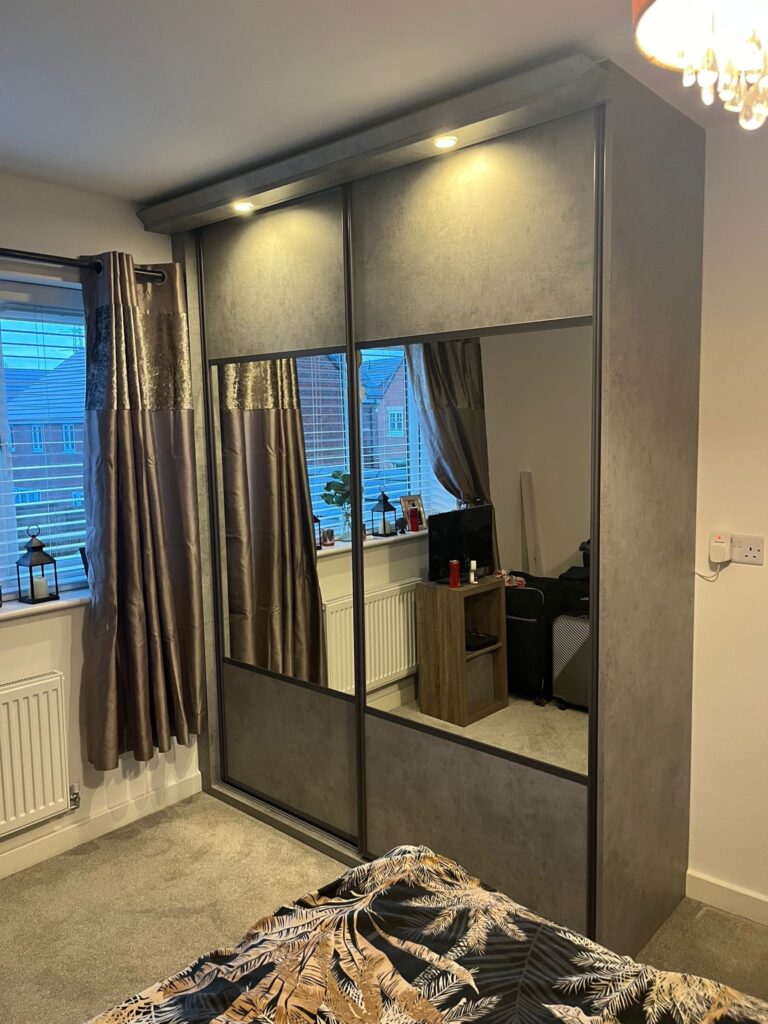 Bespoke fitted sliding door wardrobes fitted with lights above. Both doors are finished with graphite frames and split with a concrete coloured panel at the top and bottom and a large mirror in the centre