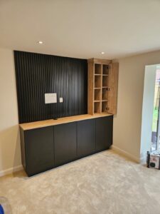 Media wall fitted with black acoustic wall paneling. There are cupboards fitted along the full width of the media wall in the colour black with a Halifax Oak top seperating the cupboards and the acoustic panelling. On the right hand side, above the cupboards, a column of Halifax Oak open shelving is fitted followed by a Halifax Oak cupabord with shelving. The cupboard door is open.