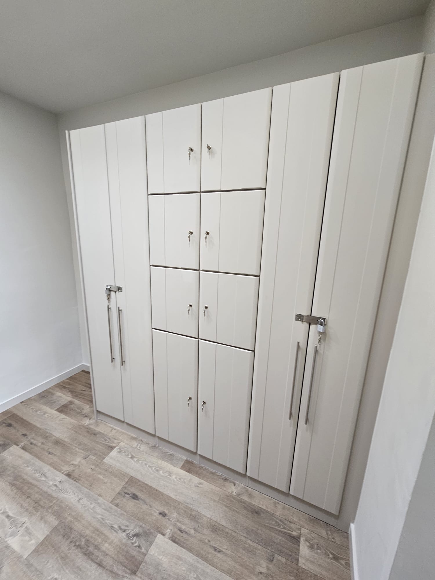 Cashmere coloured lockers manufactured to look like furniture. The doors are in the 'Grove' style. They have a wide verticle groove down the centre of the door. There are tall double lockers on the left and right -hand sides. The bottom third of the entre is fitted with a double locker. The remaining two thirds are fitted with small double lockers.