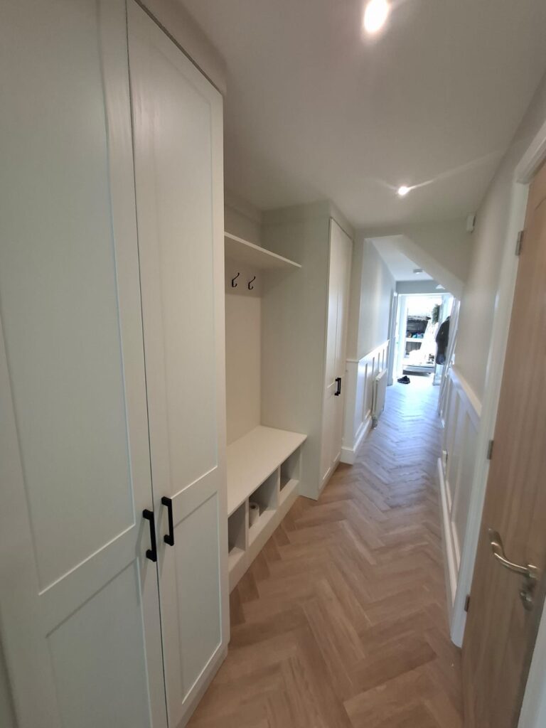 fitted bespoke hallway storage with two double floor to ceiling cupboards. In between is a bench with cuby holes below, a top storage shelf and coat ahngers. The doors are in the SHaker style and the colour finish is white. Matt Black Slim D style handles are fitted.