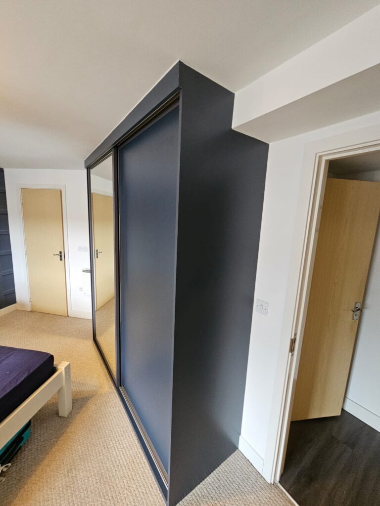Bespoke Navy sliding door wardrobes scribed around a beam at the back of the wardrobe. There are two sliding doors. One is fitted with a floor to ceiling mirror, the other a solid Indigo Blue (Navy) panel.