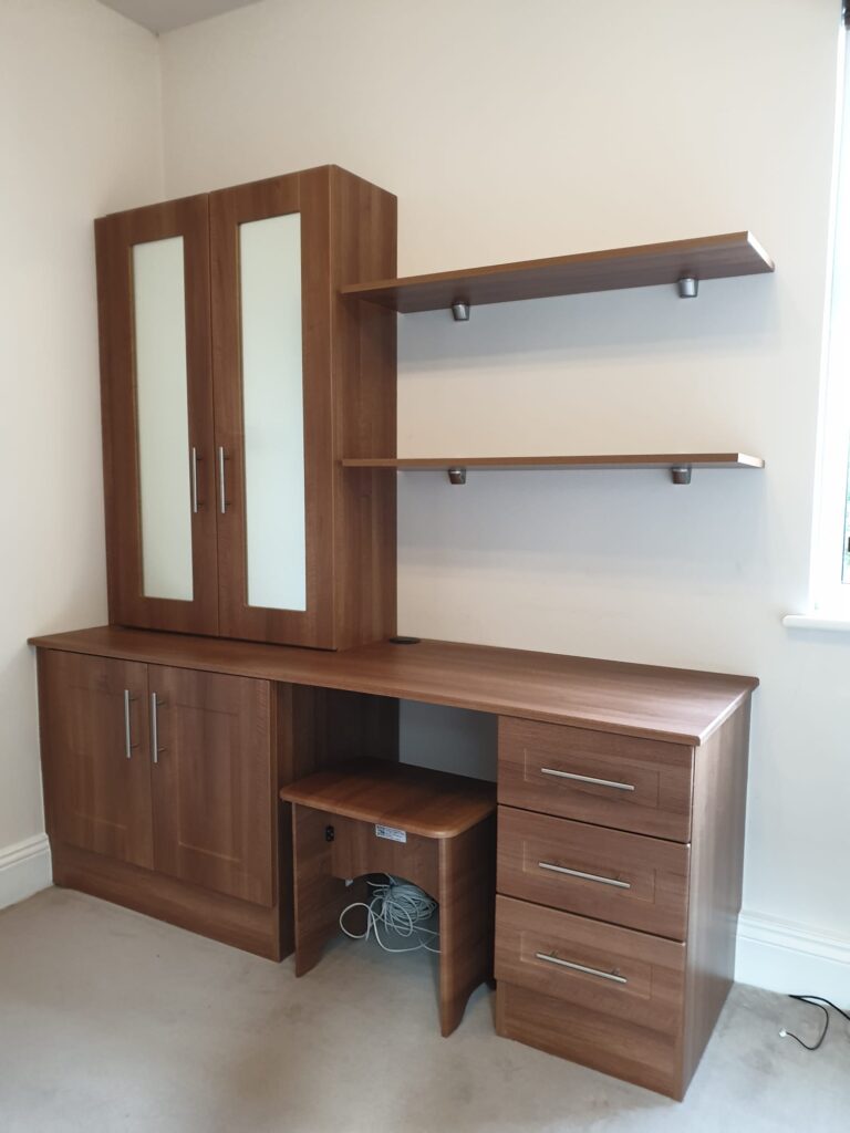 Fitted bespoke office furniture with a low unit cupboard on the left, a sitting space in the centre and four drawers on the right. There is a stool in the knee space. On the left hand side on top of the low cupboard is a shallow cupboard with frosted glass in teh doors. To the right of this are two floating shelves. Everything is in the colour Opera Walnut. The doors and drawers are in the Shaker style.