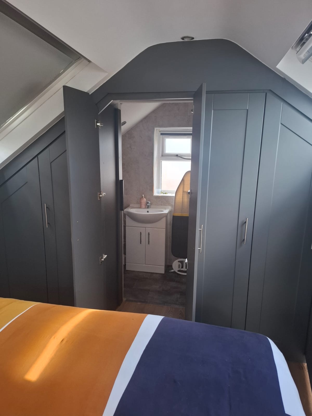 bespoke angled Shaker Grey wardrobes fitted into an apex in a loft room. Open to reveal bathroom