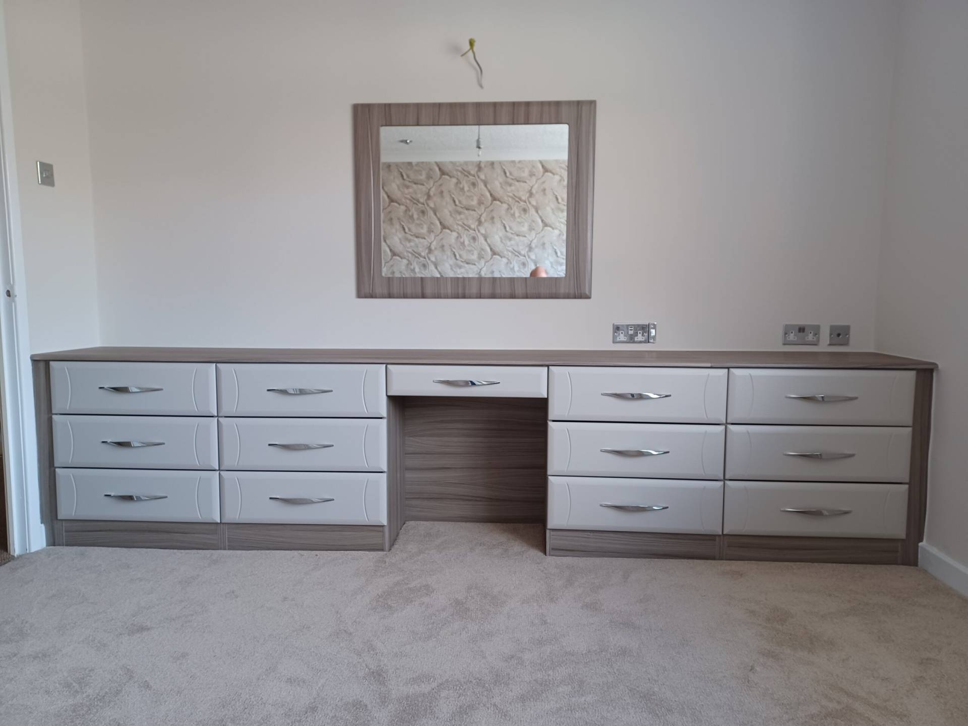 Dressing table fitted with a fitting space with a drawer in the centre. On the left and right-hand sides two sets of three drawers are fitted. The cracass is in a brown wood with grey undertones called Driftwood. The drawers are in the colour Cashmere - a neutral light grey colour with pink undertones. There is a wall mounted mirror fitted above the drawers centrally. It is Framed in the colour Driftwood to match. The mirror and desk drawer have a beveled edge all the way around. The drawers either side of the desk have a bevelved edge with a curved vertical groove on the left and right hand sides. Chrome bar handles are fitted. 