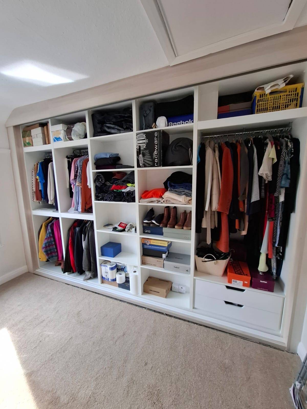 Bespoke fitted wardrobe interior with a mixture of shelving, hanging space and drawers. There is some narror shelvng fitted over a chimney breast which has been disguised by the fittings. The wardrobe is filled with clothing, shoes and boxes